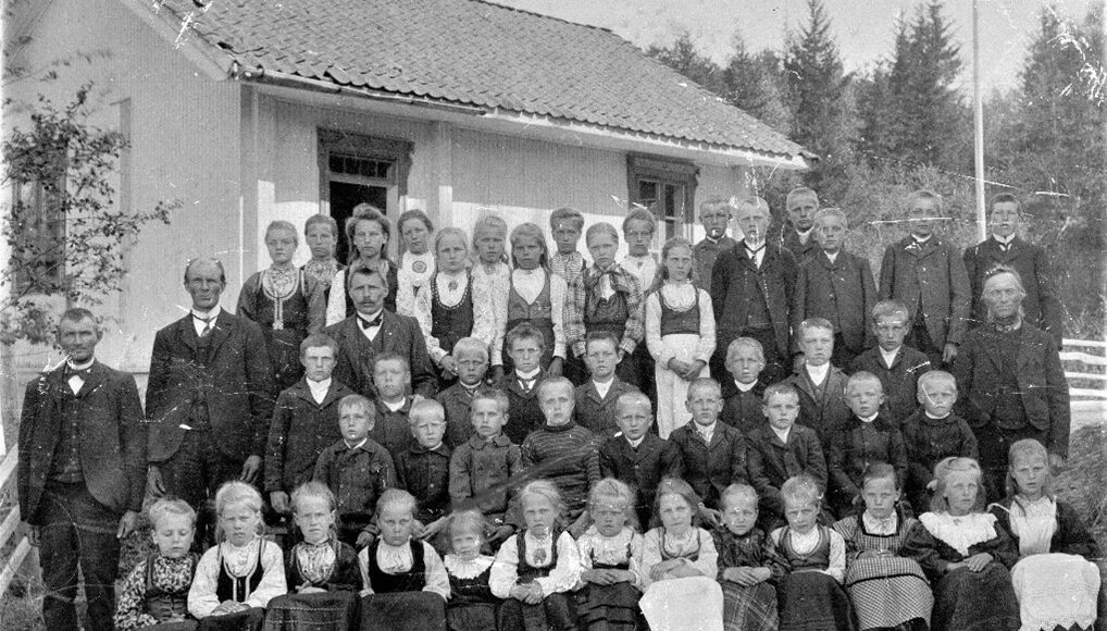 Bildet viser den eldste Strand skule, som lå på nedsiden av Søndre Vestsidaveg, (rett før avkjøringen til Søndre Strand). Bildet er tatt rundt 1907/08. Midt i elevflokken står lærer Røysland (med sløyfe). Et nytt skolehus ble bygd i 1912. Det ble brent ned i en kontrollert brann 26. april 2018. (Fotograf: Ukjent).